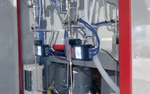 A box on the back of a fuel cell test stand has a red trim and includes metal piping, wires, and Alicat controllers.